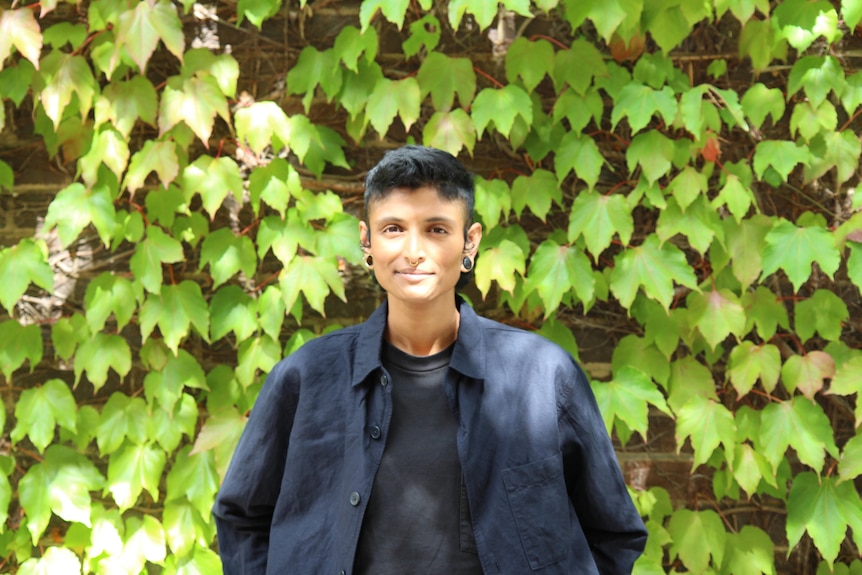 Priya Kunjan - a person with short dark hair and dark blue shirt standing in front of an ivy-covered wall
