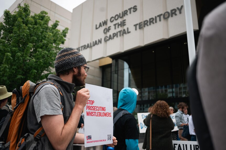 A man wearing a beanie holding a sign that says 'Stop prosecuting whistleblowers' outside an ACT law court.