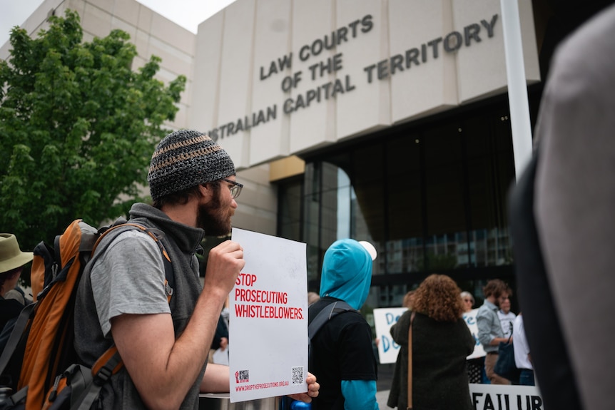 A man wearing a beanie holding a sign that says 'Stop prosecuting whistleblowers' outside an ACT law court.