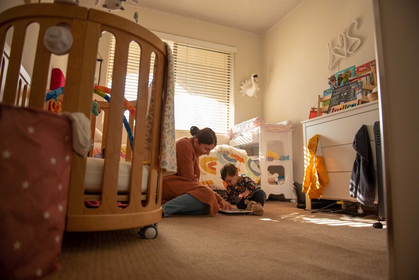 A woman plays with a baby girl on the floor of a child's room, filled with toys and a crib.