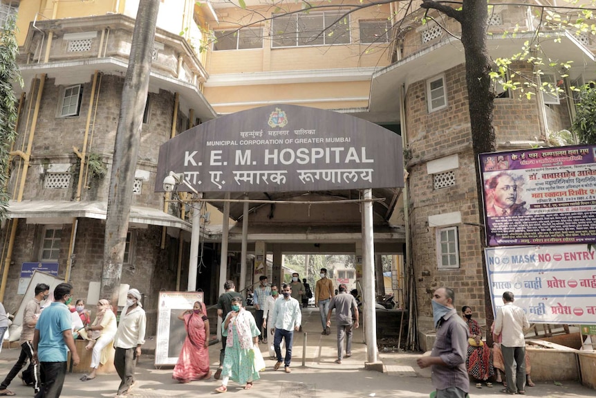 A crowd of people wearing masks walk below a sign that says KEM hospital.