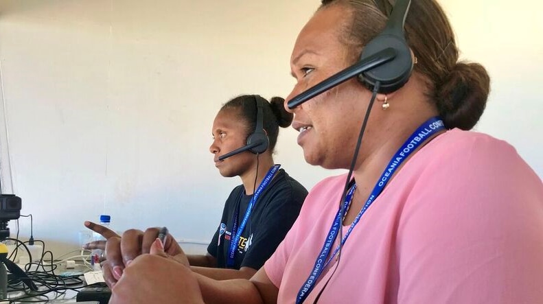 Two women with headphones in a small room look out through the window to a pitch and commentate the game.