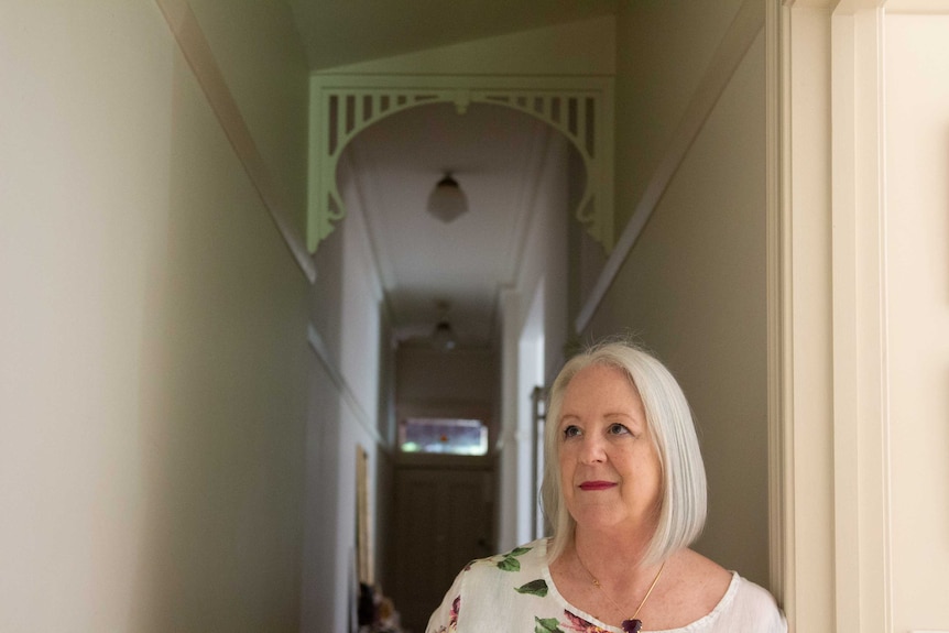 A woman with slight smile and floral print top seen from shoulders up, stands in a hall way looking up towards the ceiling.