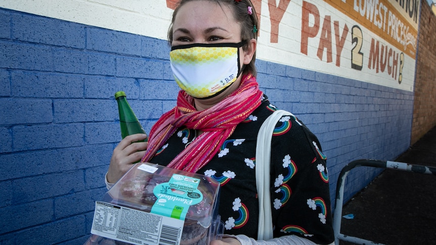 A woman wearing a mask holds a cake and drink bottle as she walks past a supermarket.