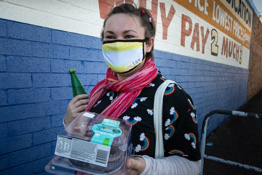 A woman wearing a mask holds a cake and drink bottle as she walks past a supermarket.