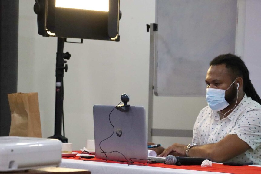 A man wearing a mask sits in an office in front of a laptop with a webcam on it and a bright studio light above.