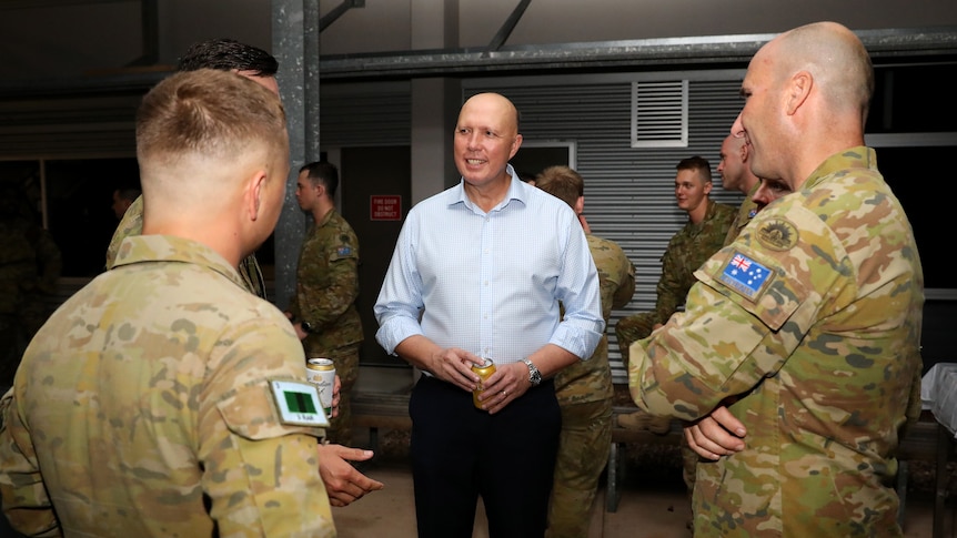 A bald man wearing a blue shirt stands talking to two soldiers dressed in army uniforms