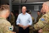 A bald man wearing a blue shirt stands talking to two soldiers dressed in army uniforms