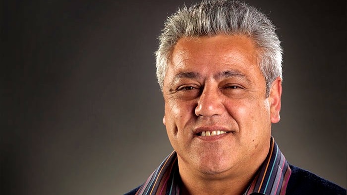 A headshot of a Maori man with short grey hair, wearing a colourful striped shirt under a suit jacket.