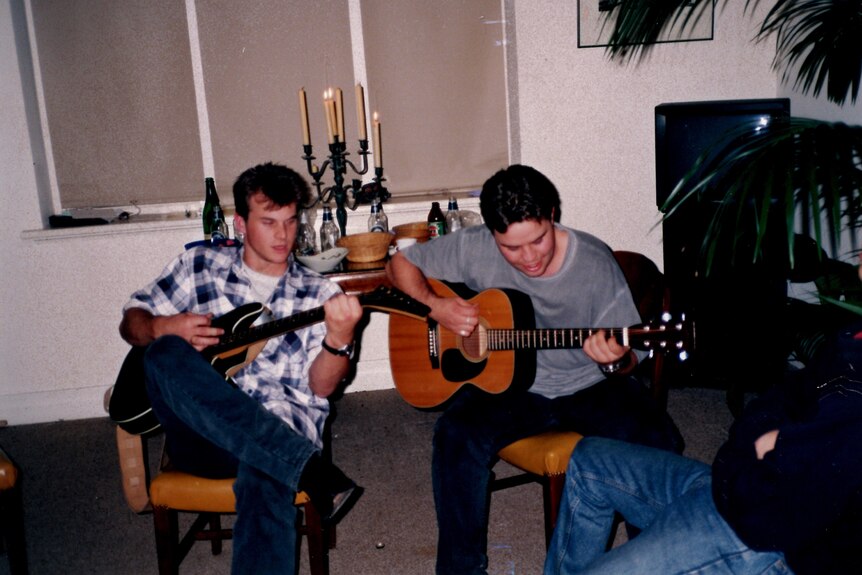 A picture of a young man looking down at the strings as he plays guitar, with another guitarist next to him.