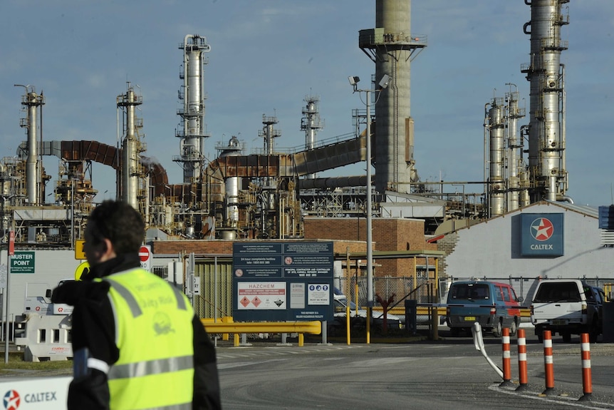 A man in fluoro yellow stands outside a gate of a large oil refinery with the word Caltex on its signage