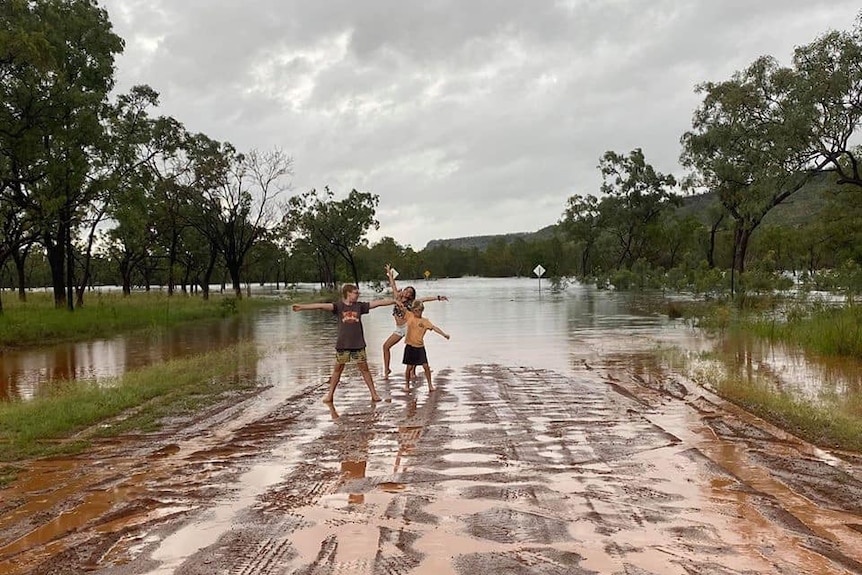 Three children posing on wet road in front of floodwaters.
