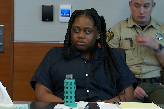 A young black girl sits at a table with a uniformed police officer behind her. 