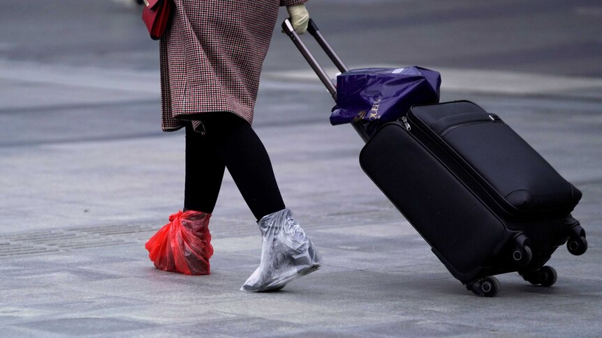A woman wheeling a suitcase with two plastic bags on her feet.