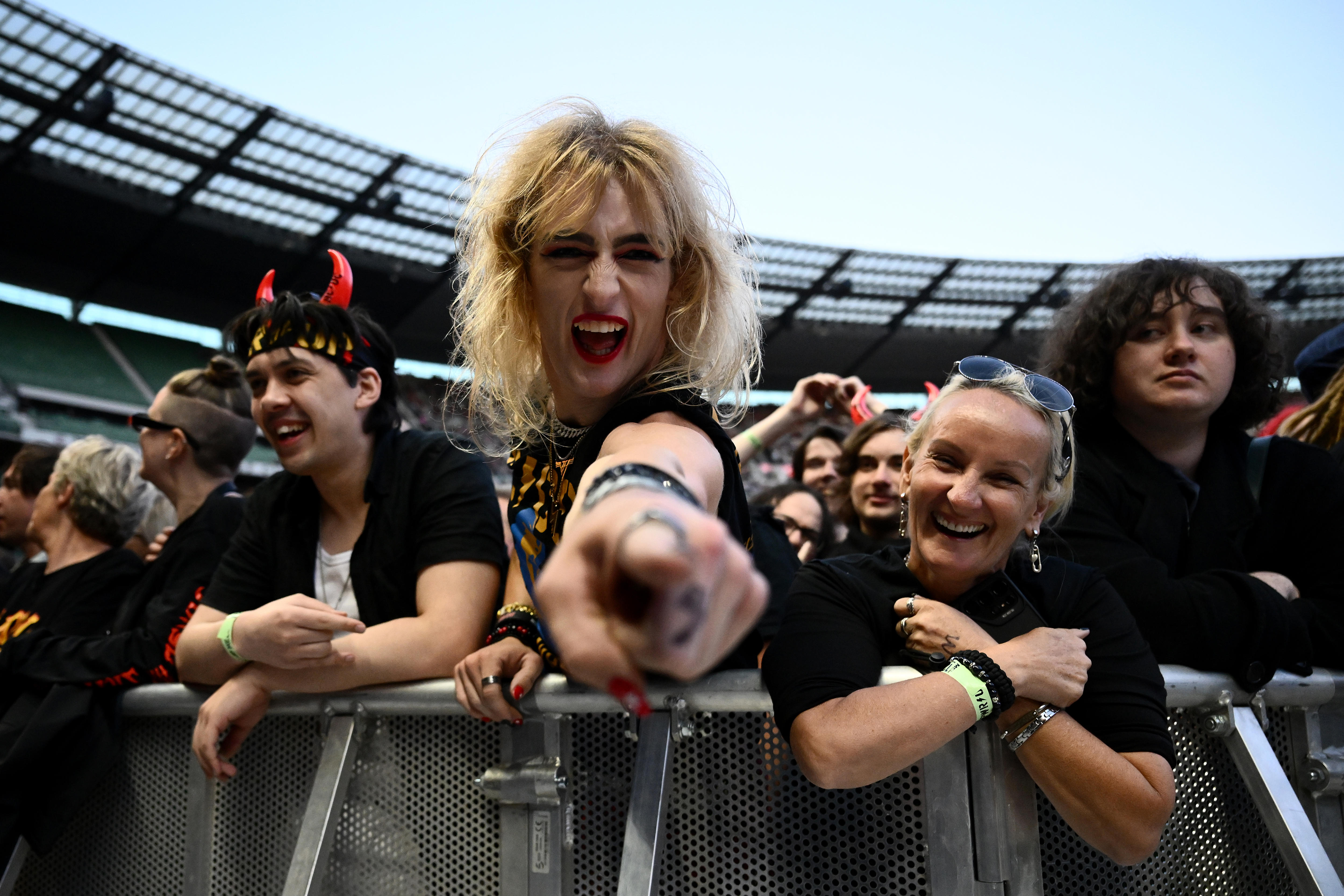 A person with long dyed blonde hair points and grins at the camera in a crowd of people in black, incuding one with devil horns.