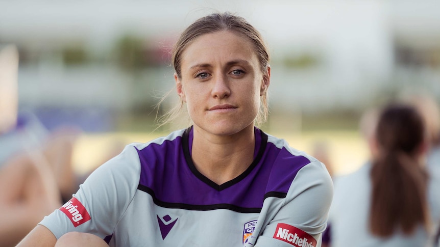 A woman wearing a sports jersey sits on the ground looking directly at the camera
