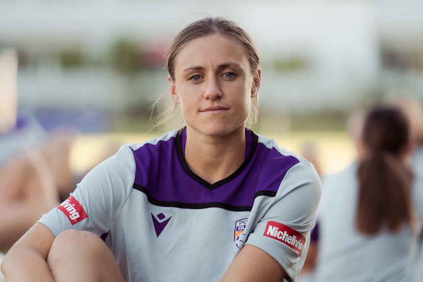 A woman wearing a sports jersey sits on the ground looking directly at the camera