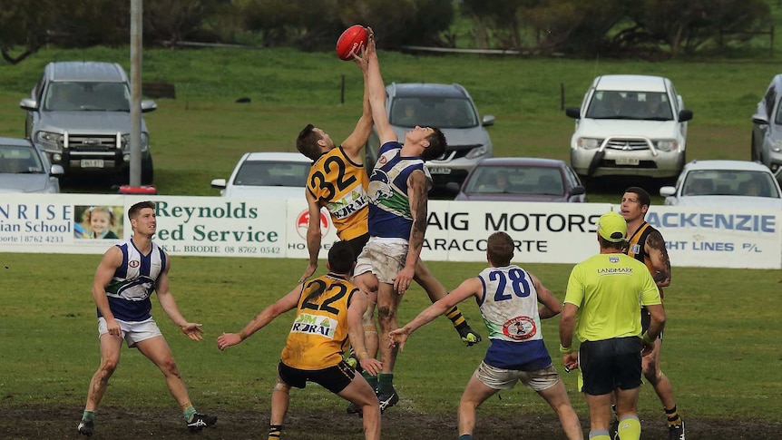 Men covered in mud and wearing football uniforms take part in a ruck contest with cars lining the oval