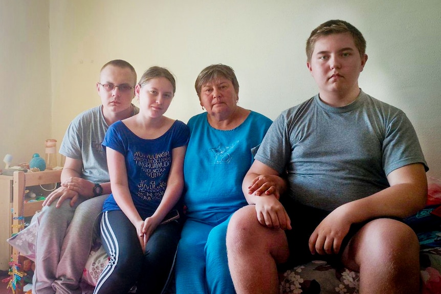 Two young boys, a young girl and a woman sit on a bed, and look at the camera with neutral expressions.