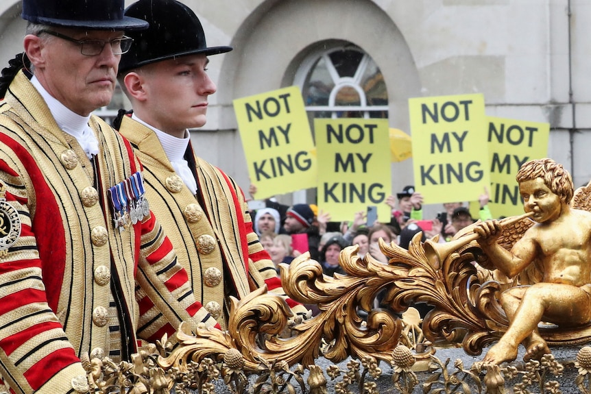 Members of the coronation procession can be seen riding past on a coach with people holding Not My King signs in the background