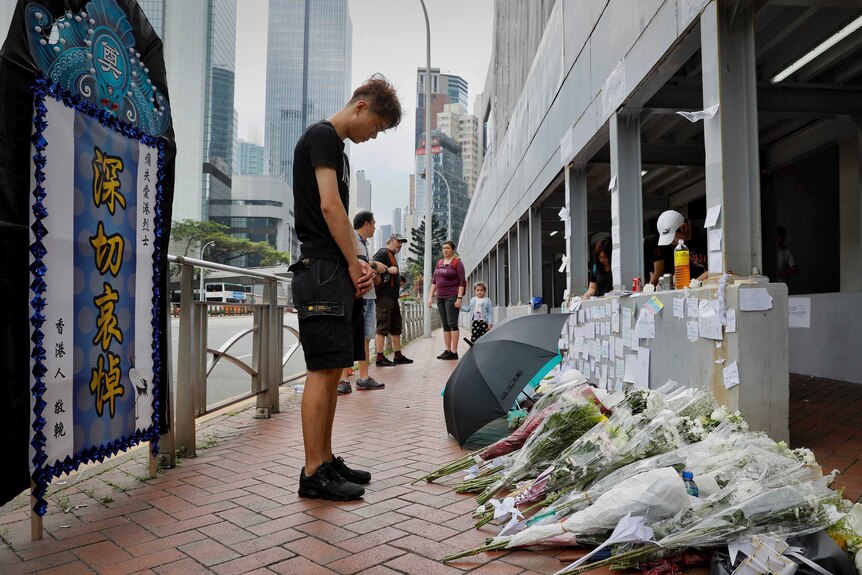 A man stands in front of a wall of flowers and messages with his head bowed.