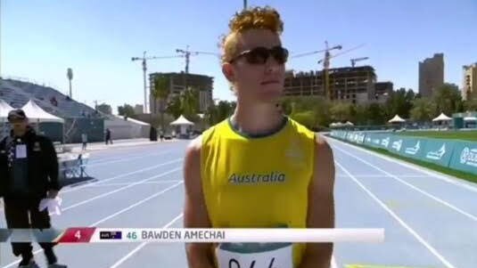 A young male runner wearing a yellow singlet with the word Australia,  and a race bib, stands on a running track.