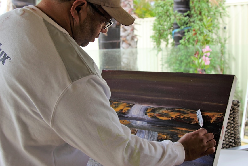 A man in a white shirt and cap paints a waterfall on a canvas 