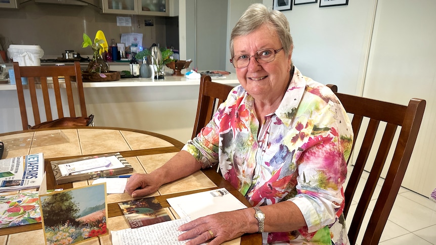 A smiling woman with grey hair and a floral shirt sits at a table with a pile of letters