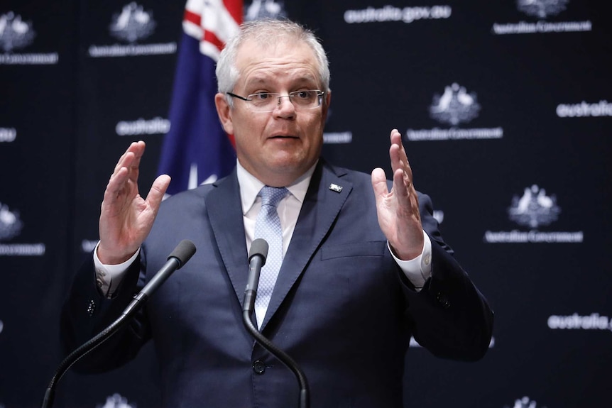 Man with grey hair and glasses in a suit at a podium with his hands raised mid-sentence.