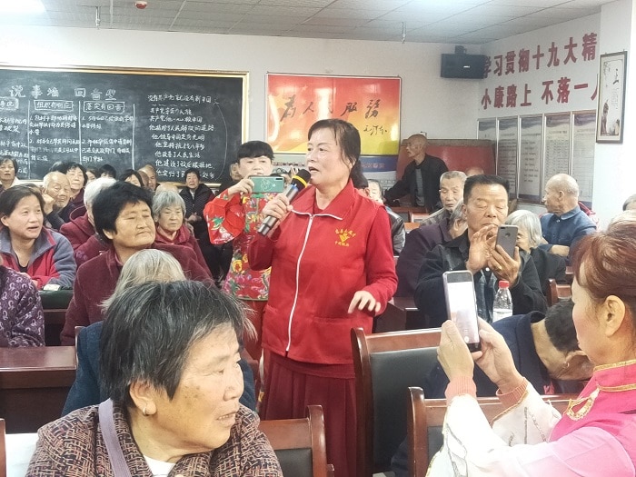 A woman in a red tracksuit sings into a microphone and elderly people watch