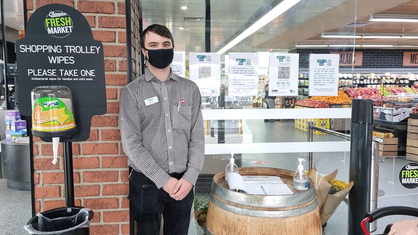 A young man wearing a mask stands at the entrance to a supermarket, with hand sanitiser on a table nearby.
