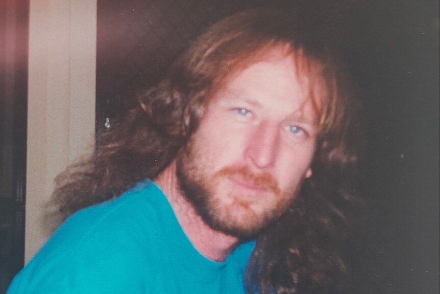 A man with long red hair and a red beard, wearing a blue t-shirt, looks at the camera with a small smile.