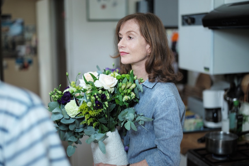 A middle aged woman with shoulder length brown hair wears chambray shirt and holds bouquet of flowers in kitchen in the daytime.