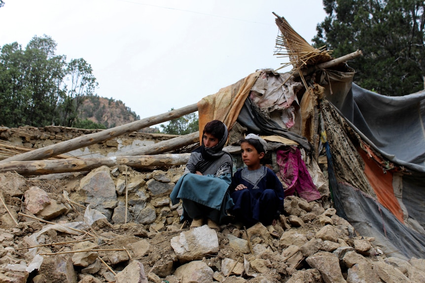 Dos niños se sientan en un montón de rocas con vigas caídas y la tela de una casa detrás de ellos 