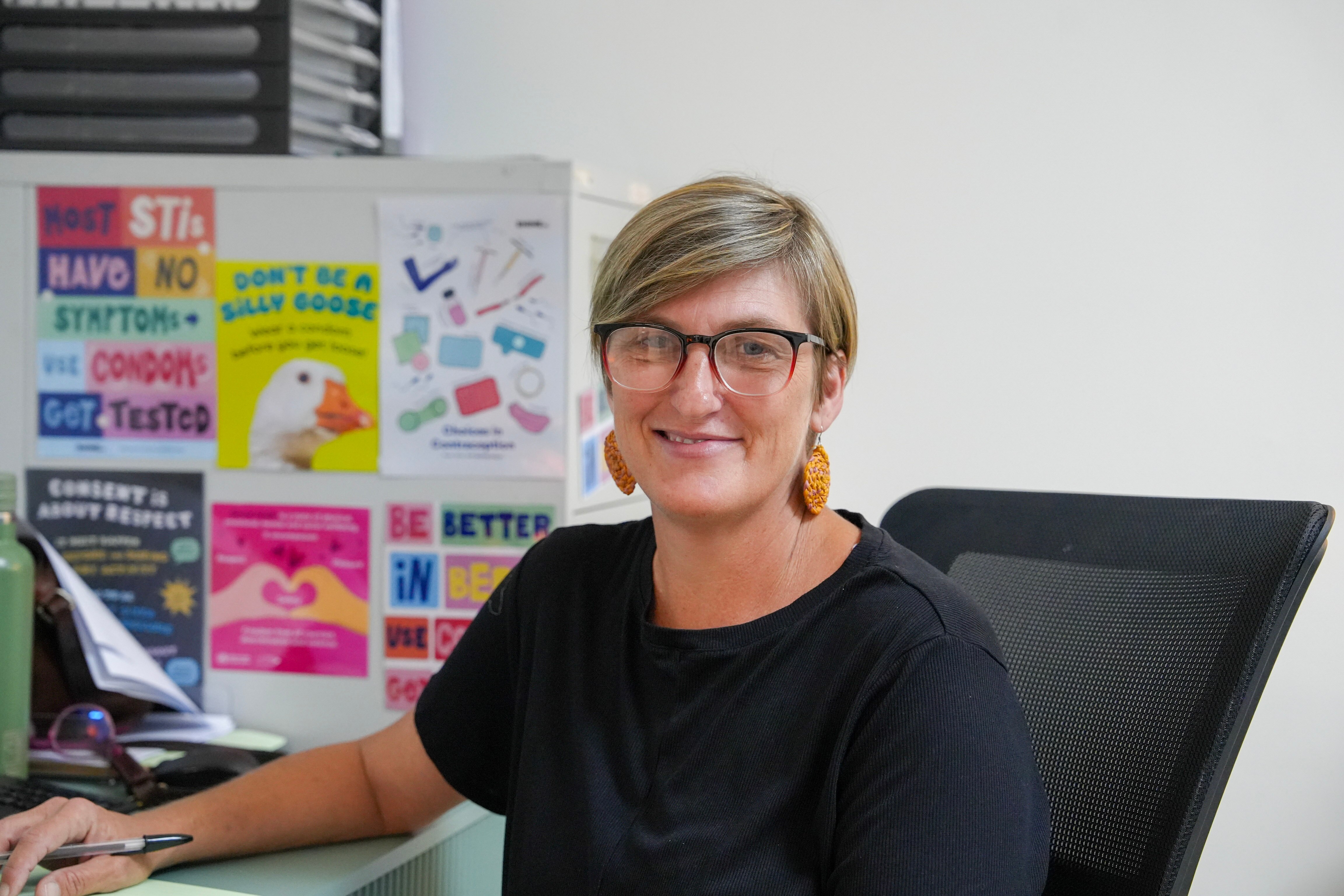 机の椅子に座っている眼鏡をかけた女性、ファイリングキャビネットの前に、性的健康に関するカラフルなポスターが付いている女性。