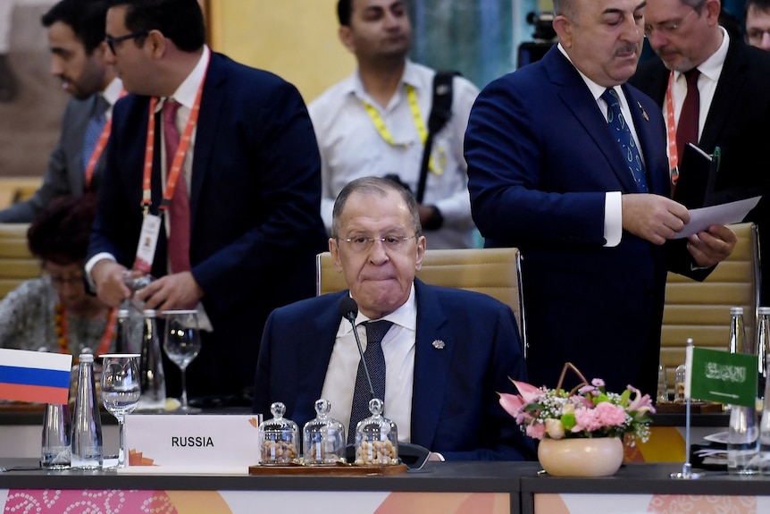 Sergei Lavrov sitting at the G20 with people walking around behind him 