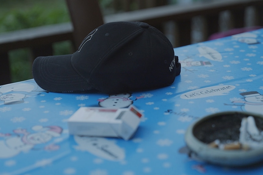 A baseball cap and packet of cigarettes on a table with a Christmas tablecloth.