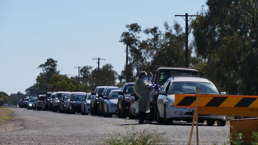 Cars line up bumper to humper along an isolated road. Two people in PPE conduct COVID-19 testing through a car window.