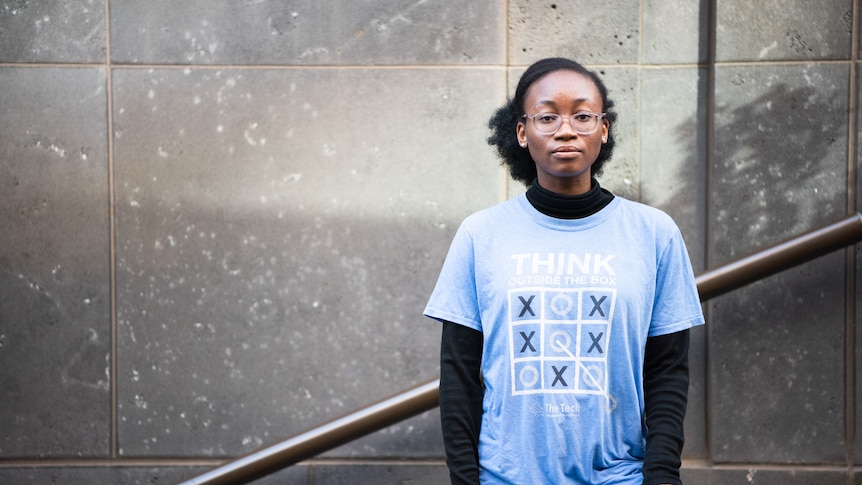 Plan youth activist Iremide Ayonrinde is standing against a wall, wearing a light blue t-shirt. 