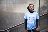 Plan youth activist Iremide Ayonrinde is standing against a wall, wearing a light blue t-shirt. 