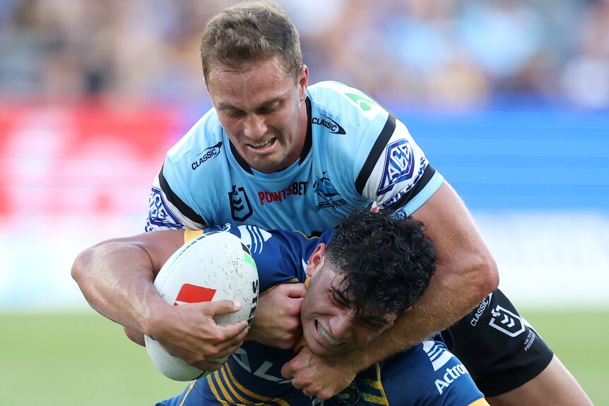 A Parramatta NRL player holds the ball as he is tackled by a Cronulla opponent.