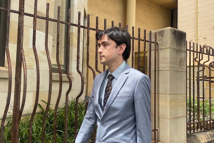 A young man in a suit walks along a footpath with a court building behind him