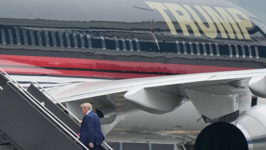 Former president Donald Trump walks up stairs to board a large aircraft with TRUMP written on the side in gold letters.