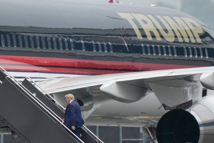 Former president Donald Trump walks up stairs to board a large aircraft with TRUMP written on the side in gold letters.