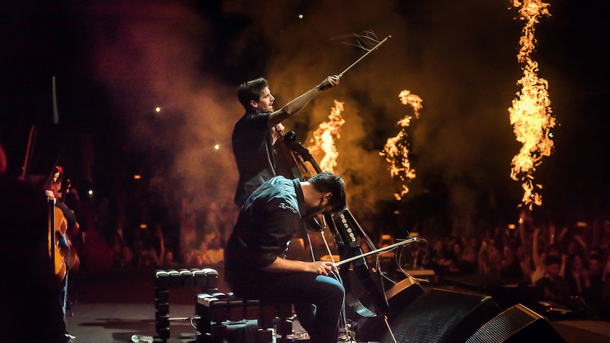 2Cellos members performing live on stage while flames shoot up around them