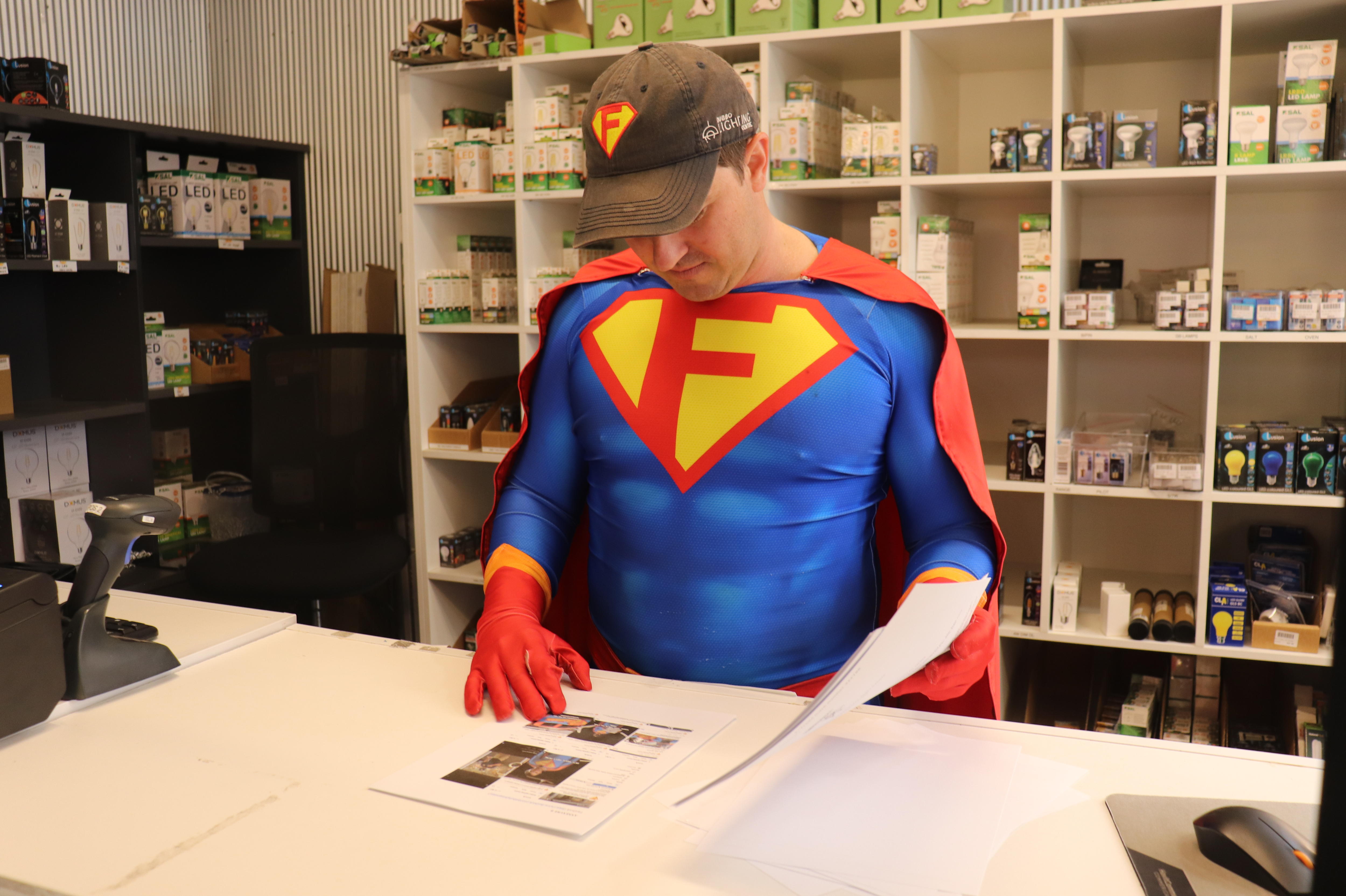 A man in a blue, red and yellow superhero costume stands looking at legal documents at a store counter.