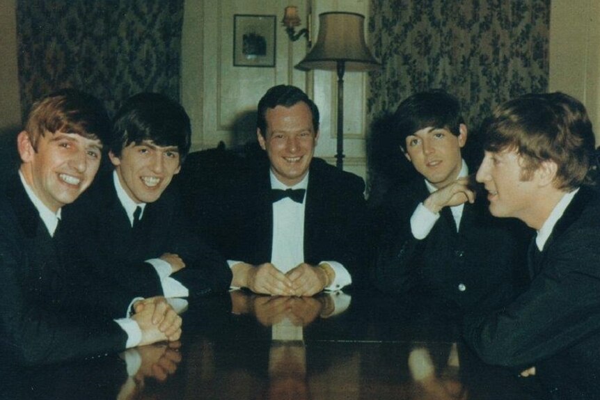 Five men in suits are sitting around a table talking and smiling