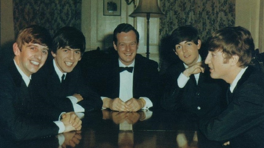 Five men in suits are sitting around a table talking and smiling