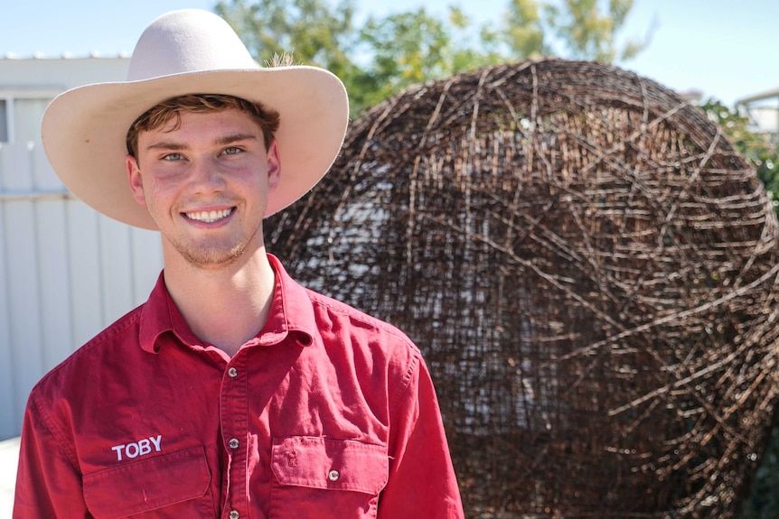 A teenager wearing a red shirt and rural cowboy-style hat stands smiling before a giant wire ball.