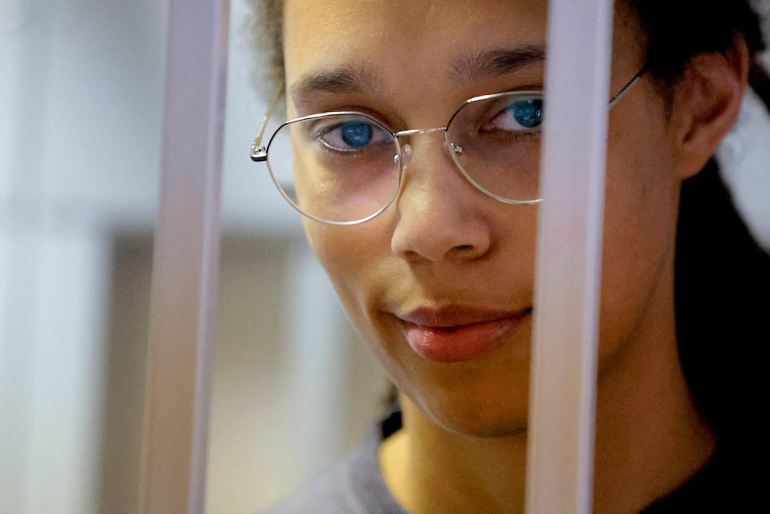 A young woman in glasses looks through prison bars